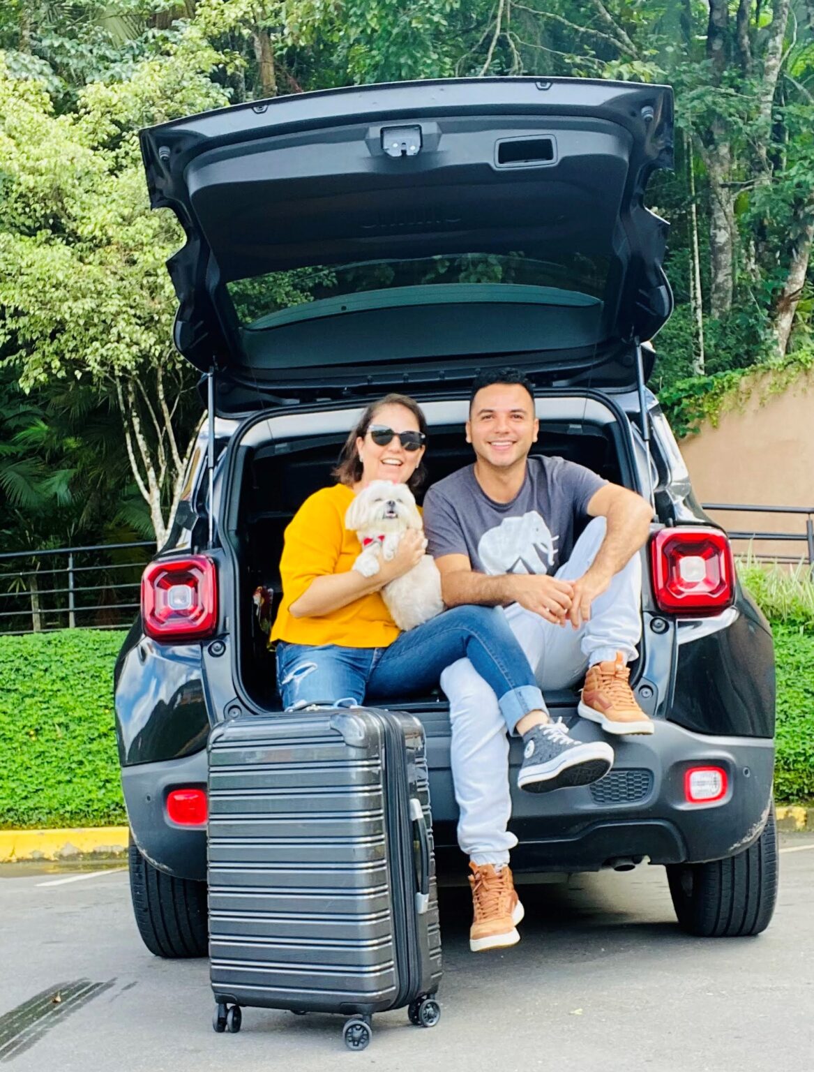 Casal sentado no porta malas do carro, segurando sua cachorrinha e com a mala de viagem na frente deles